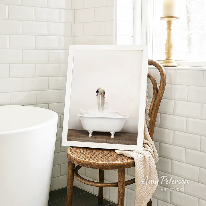 A framed photograph of a swan sitting in a bathtub is displayed on a wooden chair in a bathroom setting.