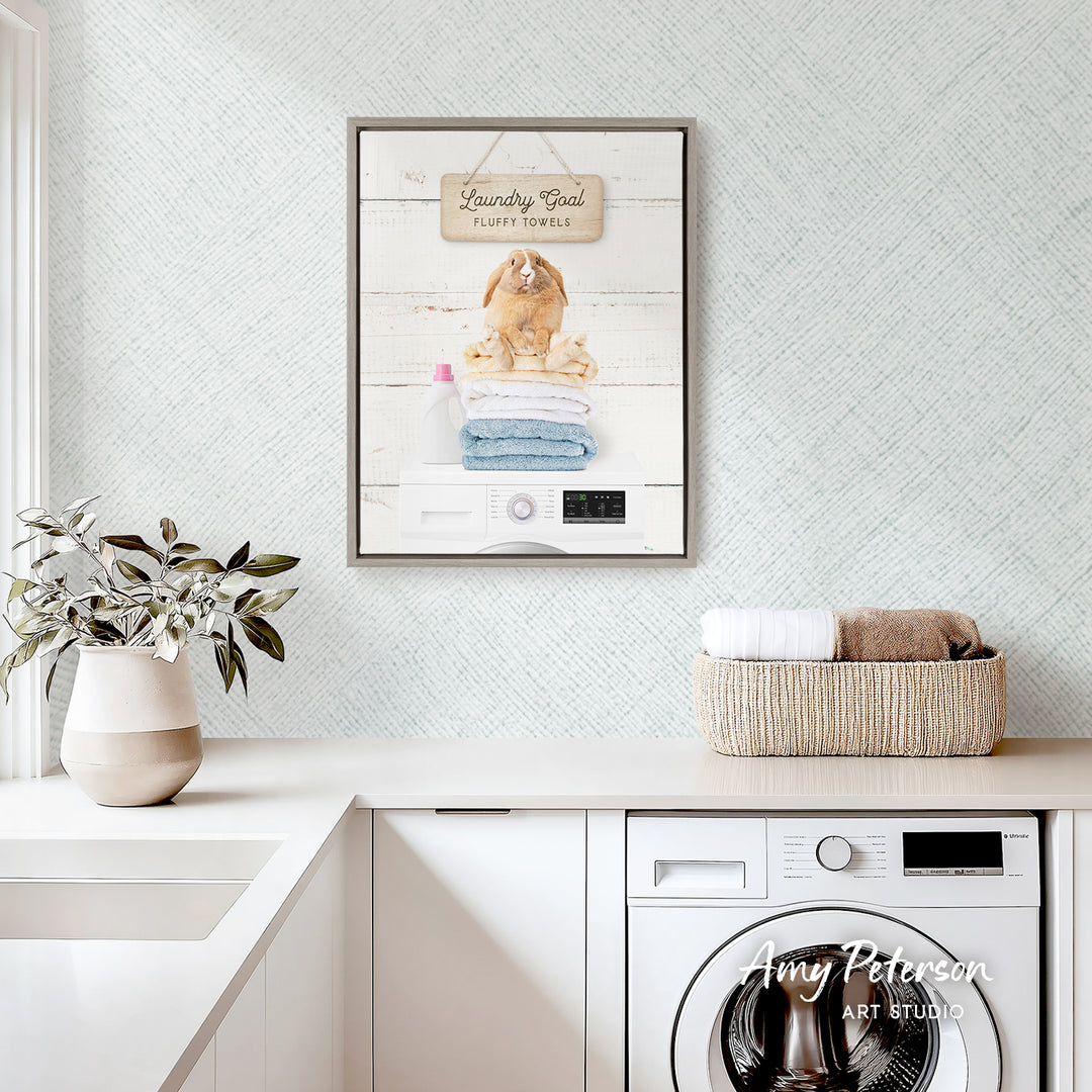 a laundry room with a framed sign that reads "Laundry Room Laundry Towels" above a washing machine. The room also includes a basket of folded towels, a potted plant, and a sink.
