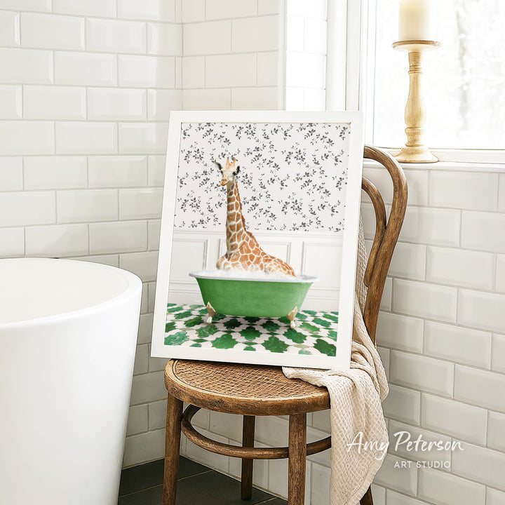 A framed image of a giraffe sitting in a green bathtub, surrounded by a patterned tile floor, is displayed on a wooden chair in a bathroom setting.