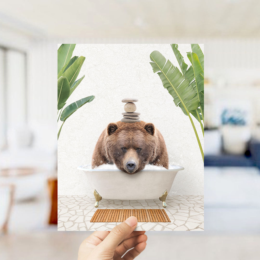 A hand holds up a framed image depicting a brown bear sitting in a bathtub filled with water, surrounded by tropical plants.