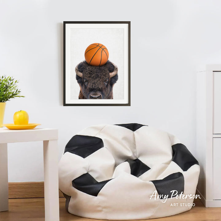 a framed picture of a buffalo with a basketball on its head, hanging on a white wall above a black and white bean bag chair.