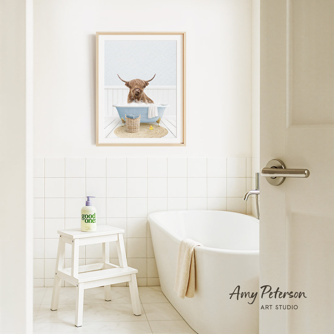 A framed image of a Highland cow sitting in a bathtub is displayed on the wall above a white bathtub in a bathroom.