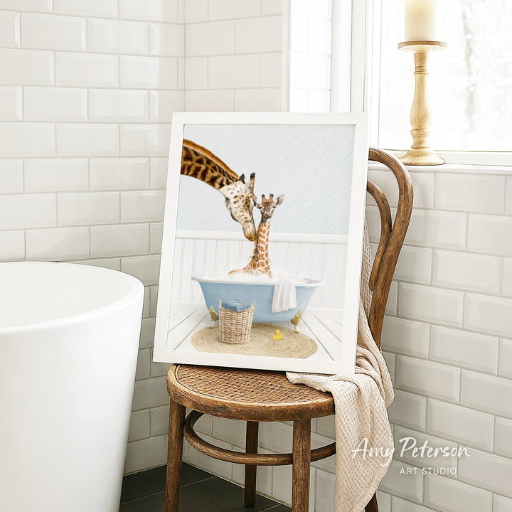 A framed image of two giraffes sitting in a bathtub, with one giraffe appearing to be bathing the other giraffe. The image is displayed on a wooden chair in a bathroom setting.