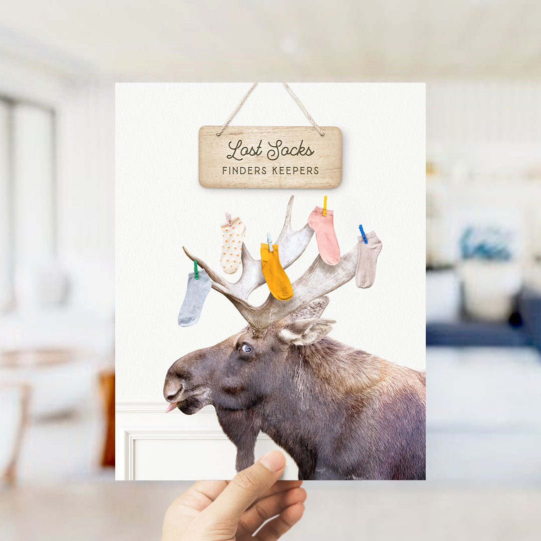 A hand is holding up a card with a moose wearing a sign that says "Lost Socks Finders Keepers".