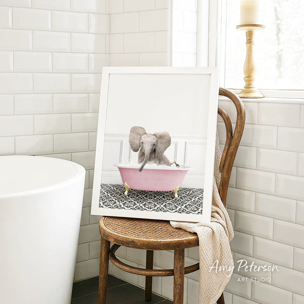 A framed image of an elephant sitting in a pink bathtub, with a towel draped over the back of a wooden chair in a bathroom setting.