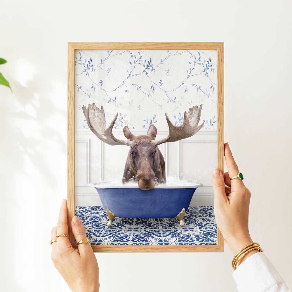 A person's hands are holding a framed picture of a moose sitting in a blue bathtub filled with bubbles, with a floral pattern on the wall behind the moose.