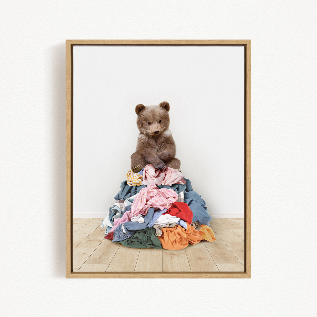 A brown bear sits atop a pile of clothes on a wooden floor.