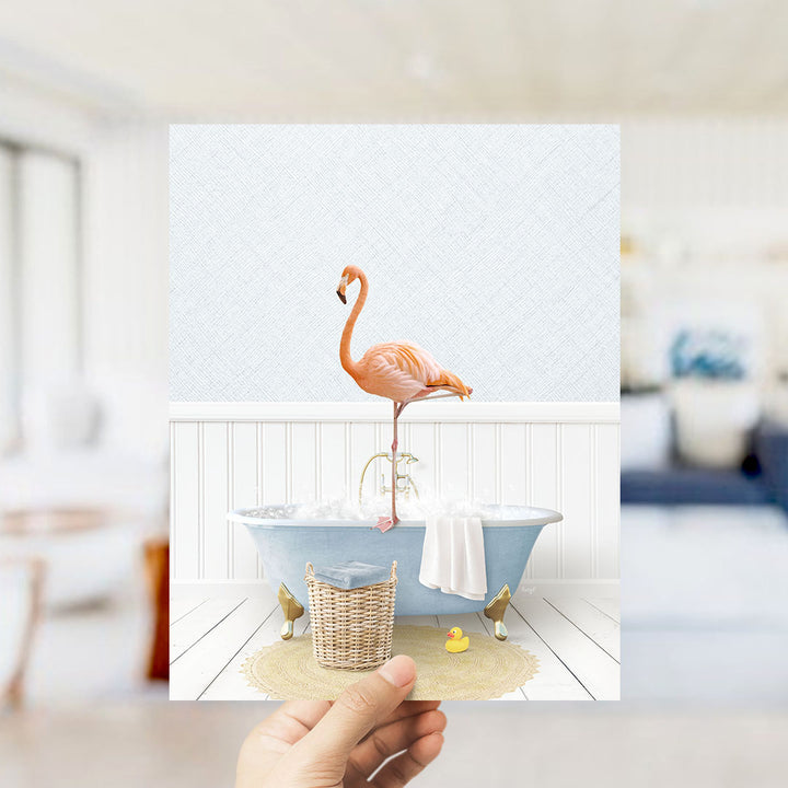 A hand holds up a framed image depicting a pink flamingo standing on a blue bathtub filled with bubbles, with a yellow rubber duck nearby.