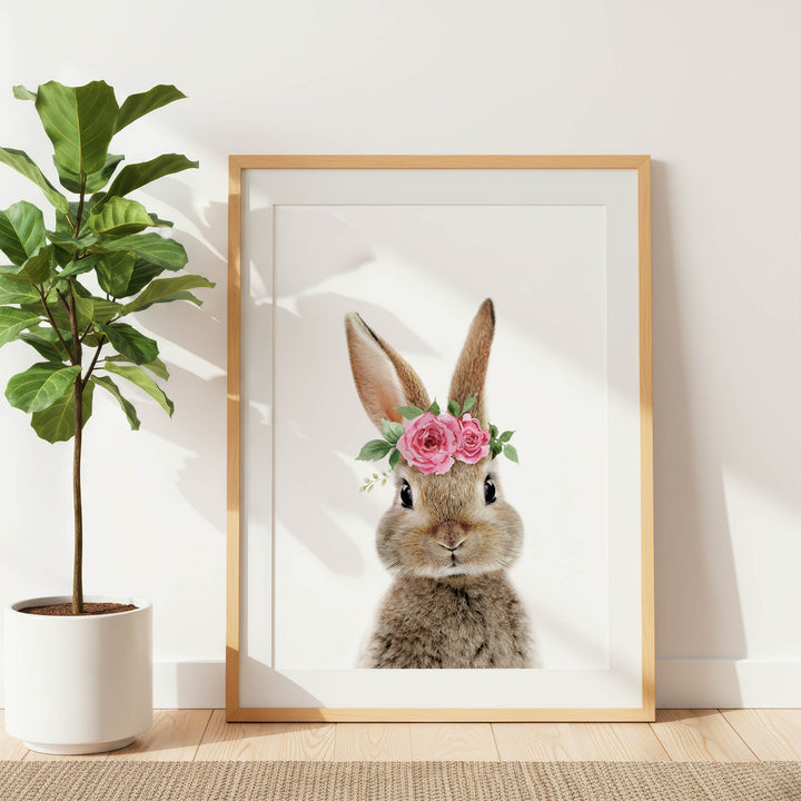 A framed picture of a rabbit wearing a pink flower crown is displayed on a white wall, with a potted plant to the left.