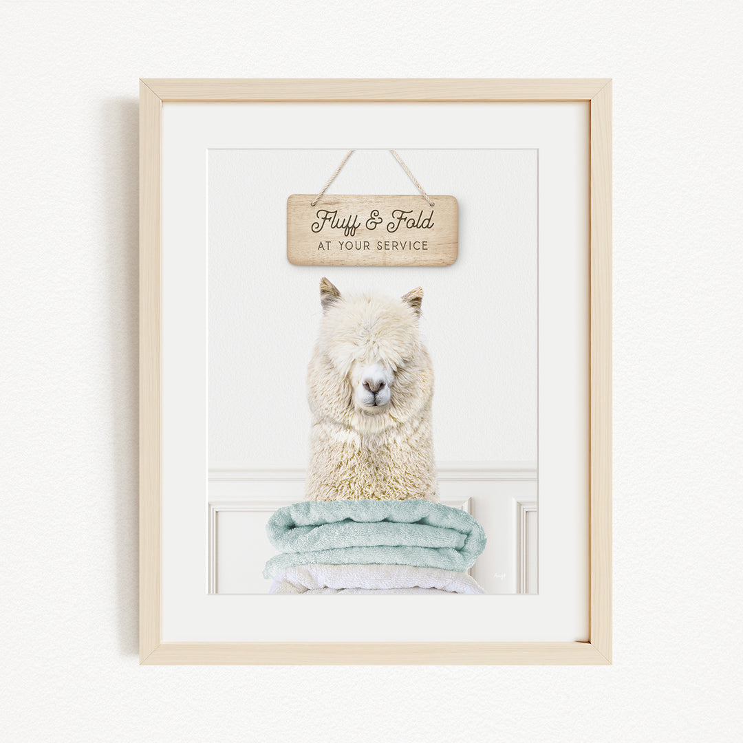 A framed image of a fluffy white alpaca sitting on a stack of light blue towels, with a wooden sign above it that says "Fluffy & Fold at your service".