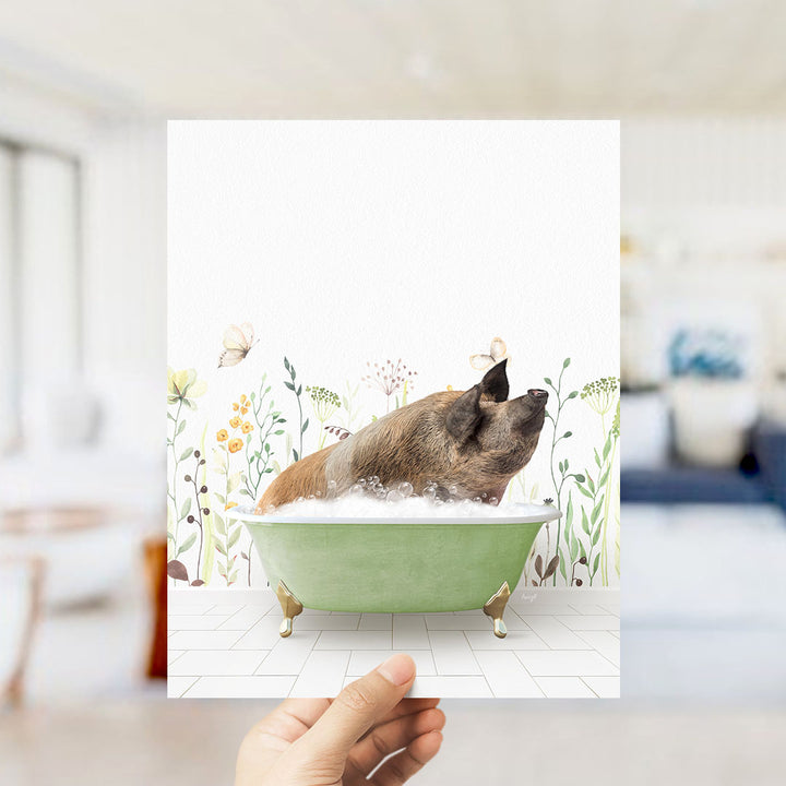 A hand holds up a framed image of a pig sitting in a green bathtub, surrounded by a floral pattern.