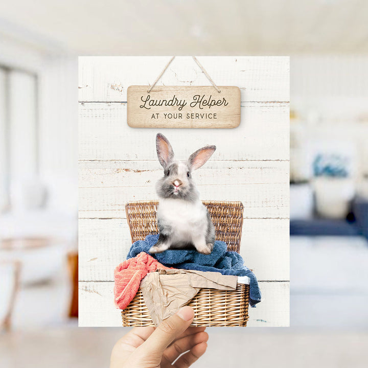 A hand holding a card with a picture of a rabbit sitting in a wicker basket filled with clothes, with a sign above it that says "Laundry Helper at your service".
