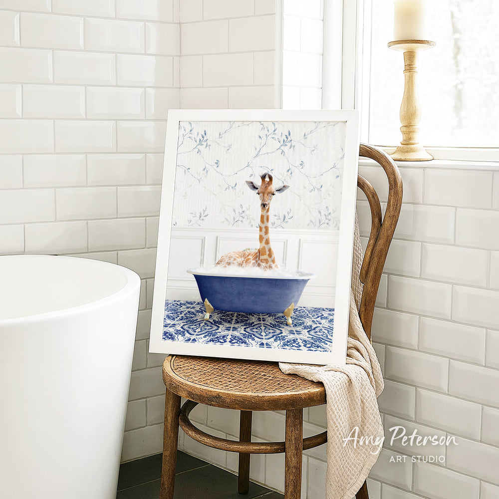 A framed image of a giraffe sitting in a blue bathtub, surrounded by a patterned tile floor, is displayed on a wooden chair in a bathroom setting.