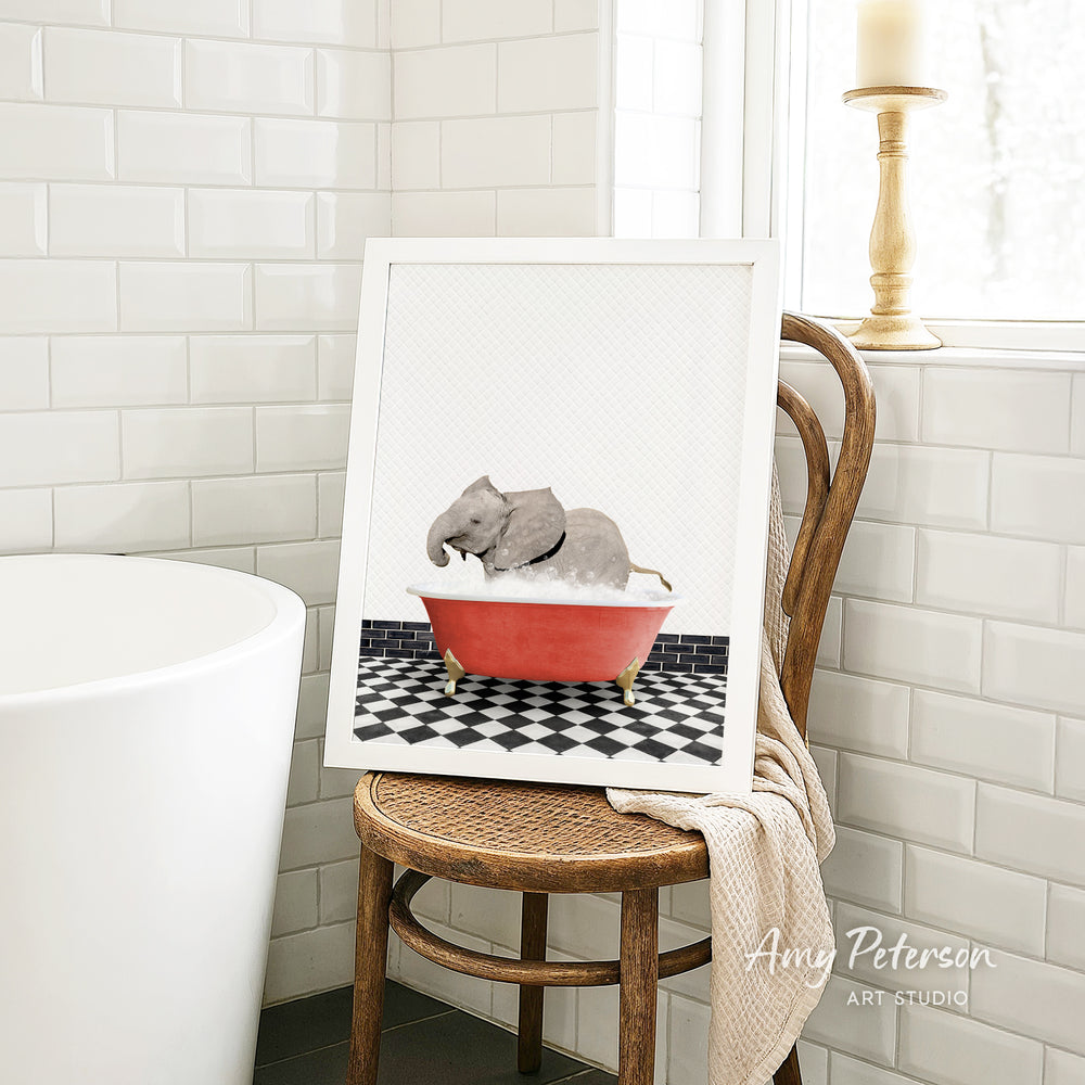 A framed image of a baby elephant sitting in a red bathtub, surrounded by a black and white checkered floor, is displayed on a wooden chair in a bathroom setting.