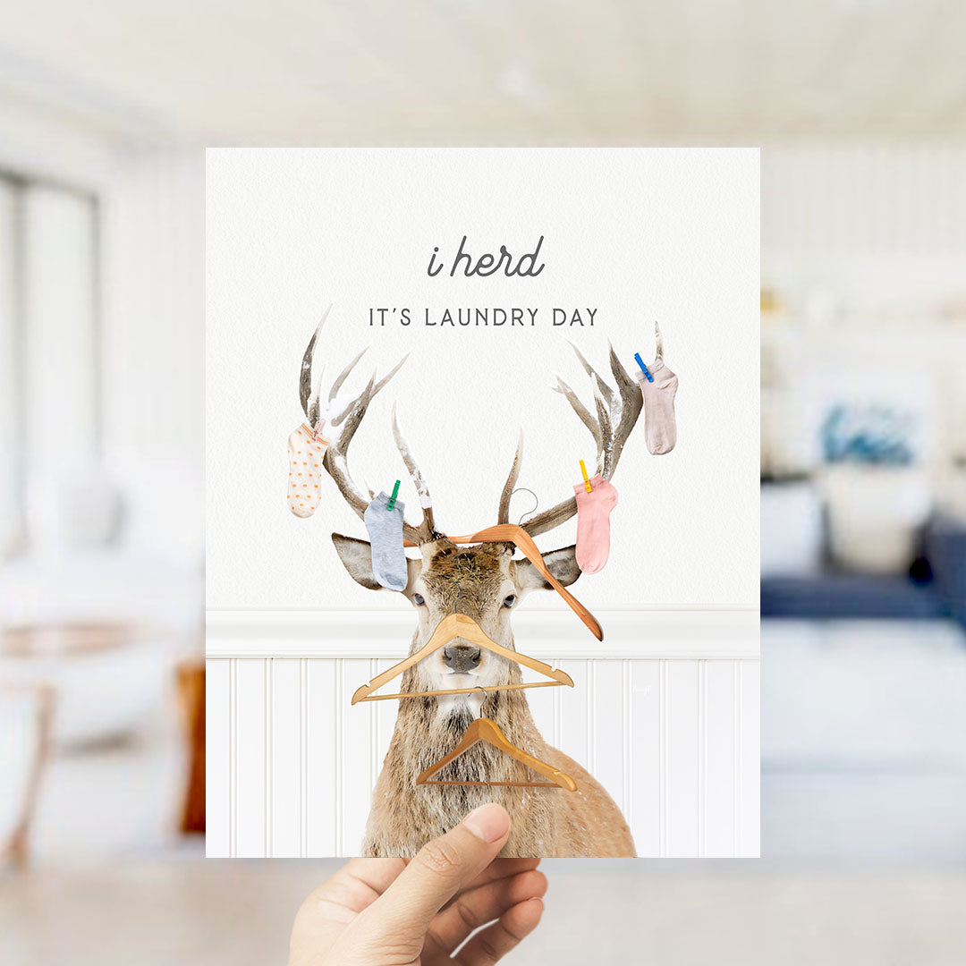 A hand holding a card with a deer wearing clothes and holding clothes hangers, with the text "It's laundry day" written on it.