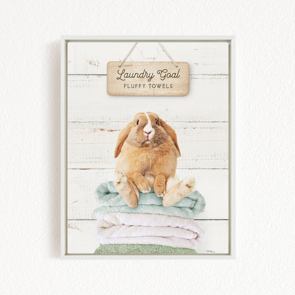 A wooden sign with the text "Laundry Goal Fluffy Towels" hangs above a stack of folded towels on a white wooden background.