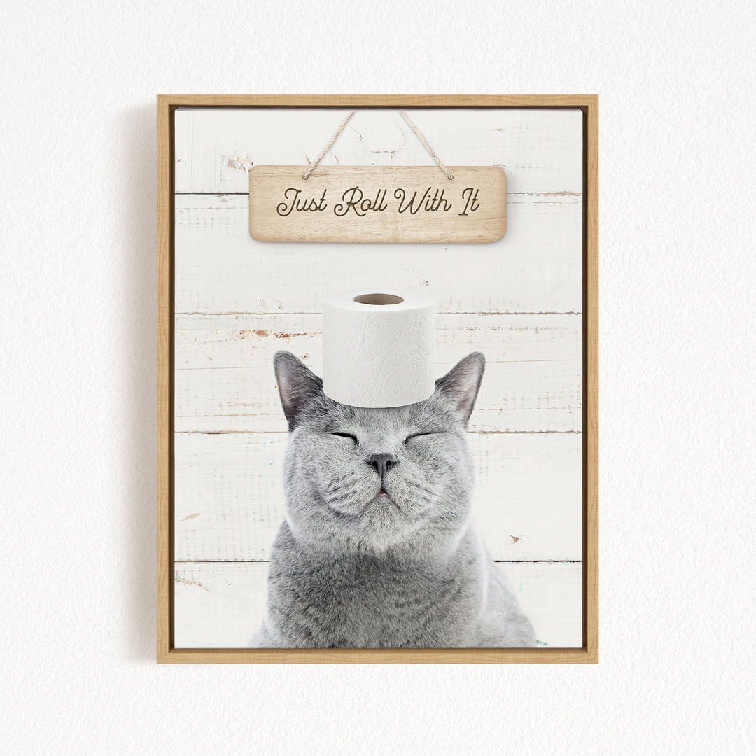 A gray cat with its eyes closed and a roll of toilet paper on its head, with a wooden sign above that says "Just Roll With It".
