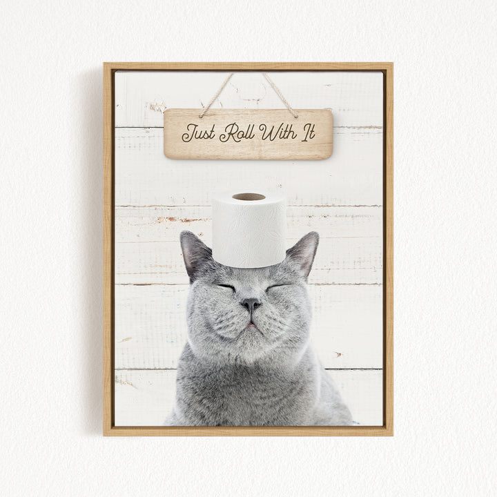 A gray cat with its eyes closed and a roll of toilet paper on its head, with a wooden sign above that says "Just Roll With It".