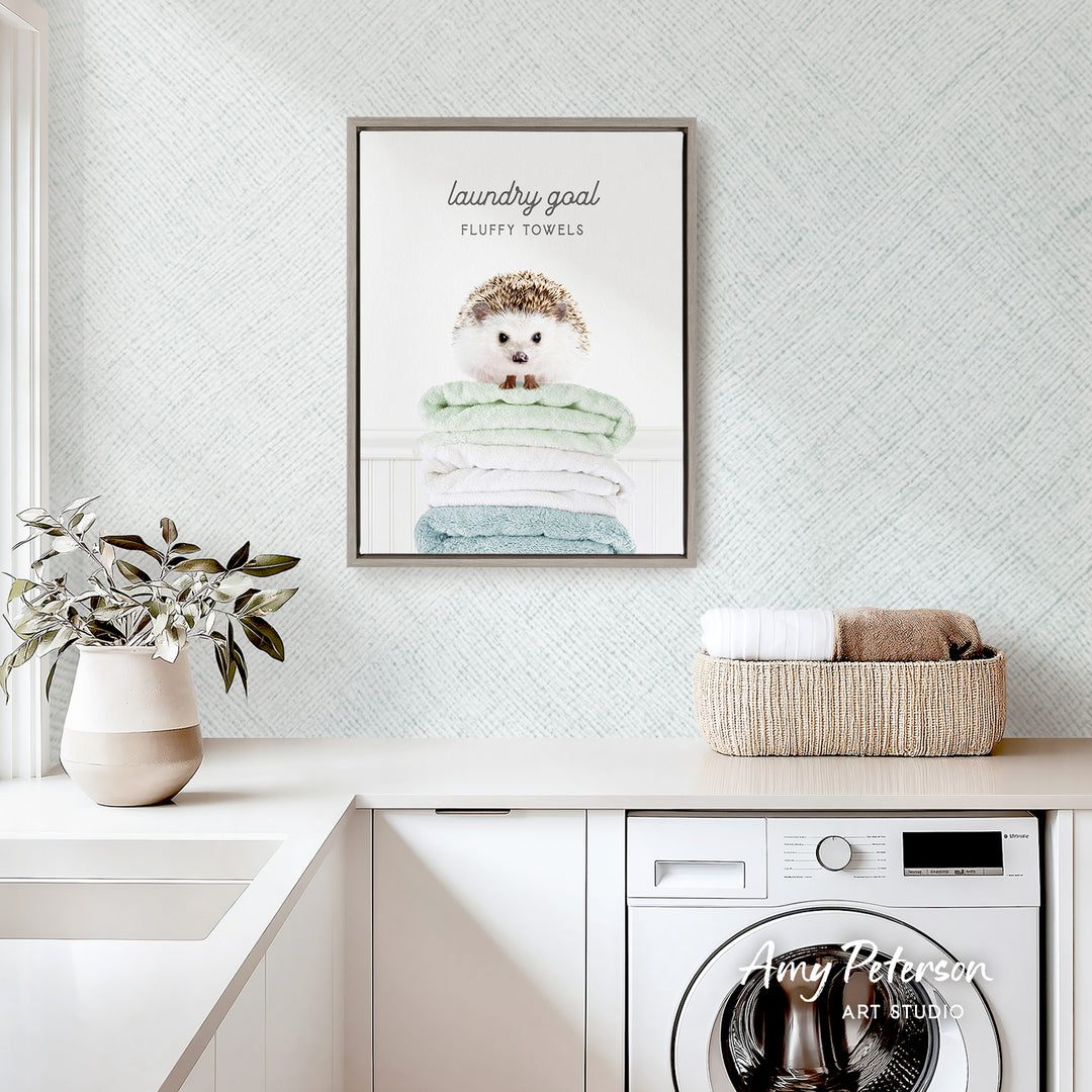 a framed poster of a hedgehog sitting atop a stack of fluffy towels, hanging on a wall above a laundry basket and a potted plant.