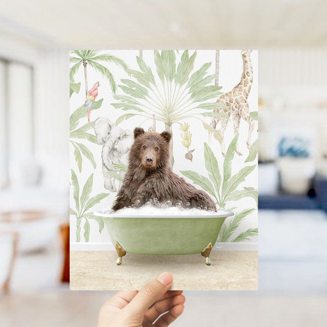 A hand holds up a framed print featuring a brown bear sitting in a green bathtub, surrounded by tropical foliage and animals.
