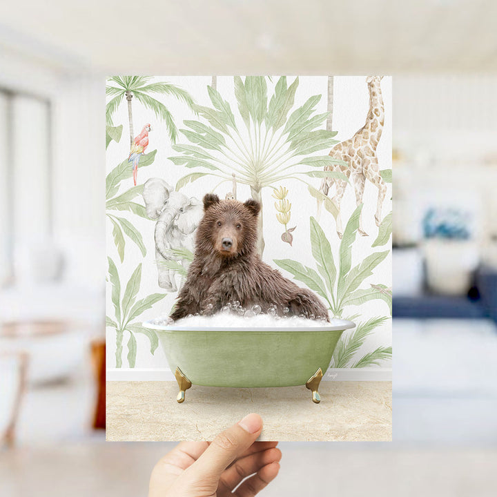A hand holds up a framed print featuring a brown bear sitting in a green bathtub, surrounded by tropical foliage and animals.