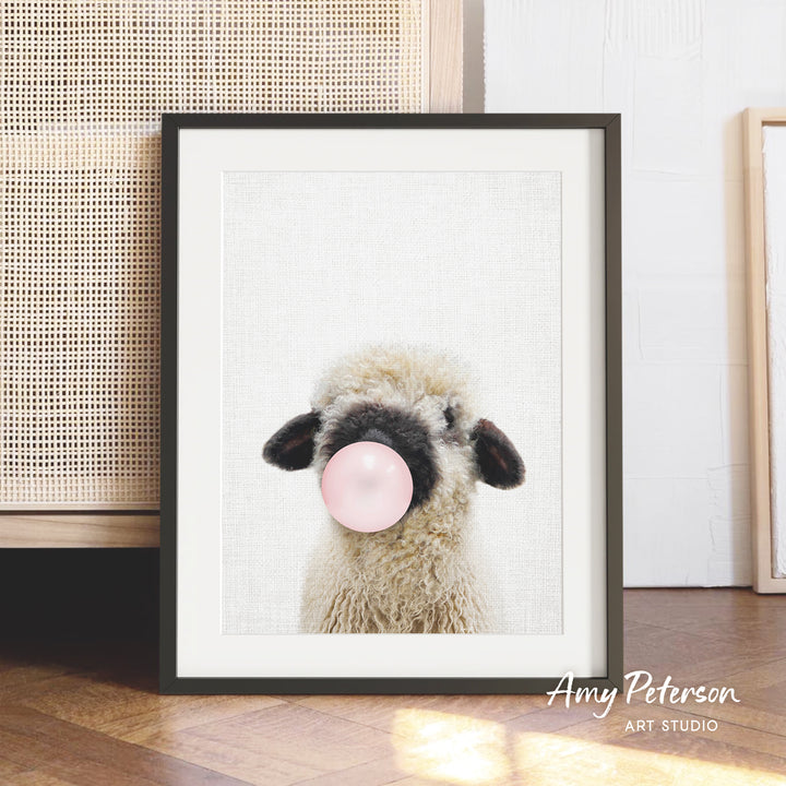 A framed photograph of a fluffy sheep blowing a bubble with a pink bubble gum bubble.