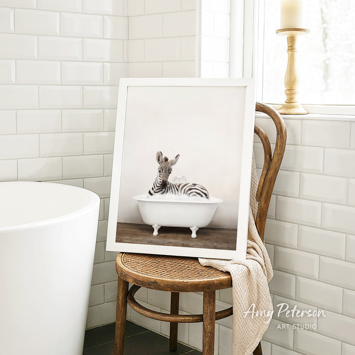 A framed photograph of a zebra sitting in a white bathtub is displayed on a wooden chair in a bathroom setting.