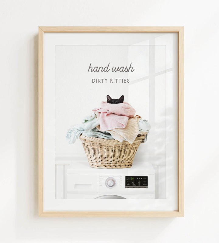 A framed image depicting a cat sitting in a basket filled with laundry, with the text "hand wash dirty kitties" written above the basket.