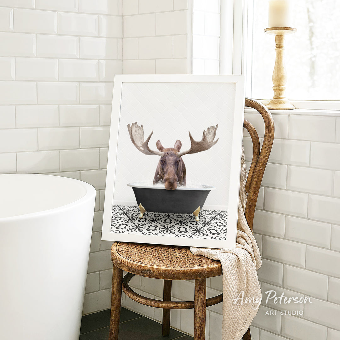A framed image of a moose with antlers sitting in a bathtub, surrounded by a patterned tile floor, is displayed on a wooden chair in a bathroom setting.