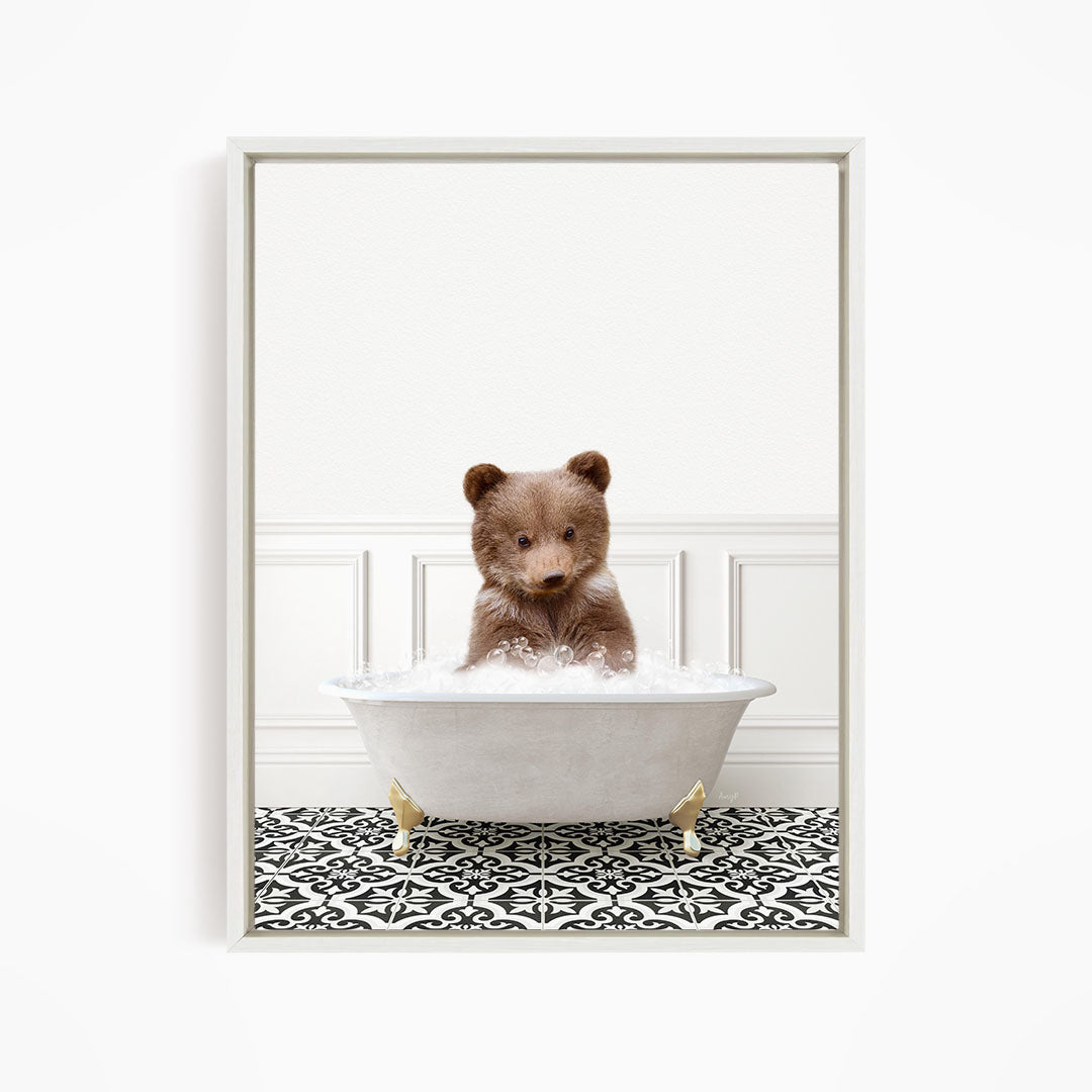 A cute brown bear sitting in a white bathtub filled with bubbles, looking directly at the camera.