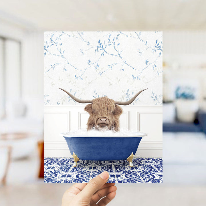 A hand holds up a framed image depicting a Highland cow with long horns sitting in a blue bathtub filled with bubbles.