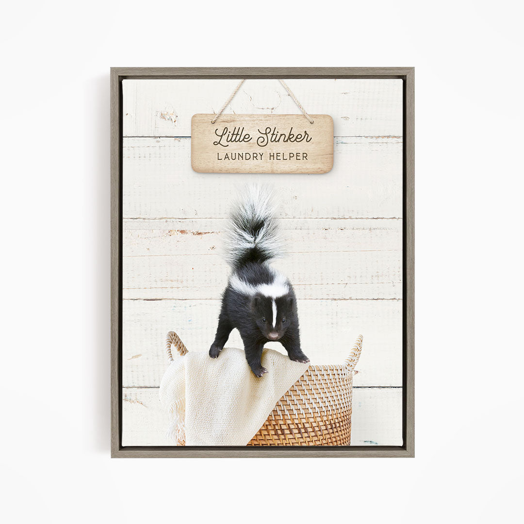 A black and white skunk standing on a woven basket, with a wooden sign above that reads "Little Stinker Laundry Helper".