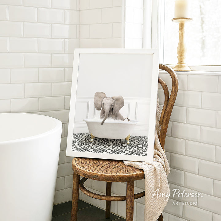 A framed photograph of a baby elephant sitting in a bathtub is displayed on a wooden chair in a bathroom setting.