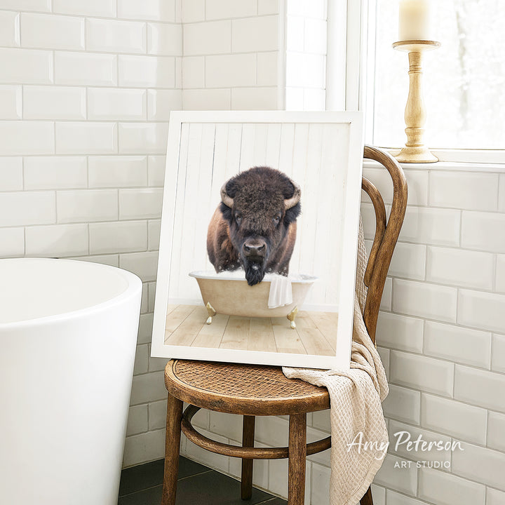 A framed image of a bison sitting in a bathtub, with a towel draped over the back of the chair.