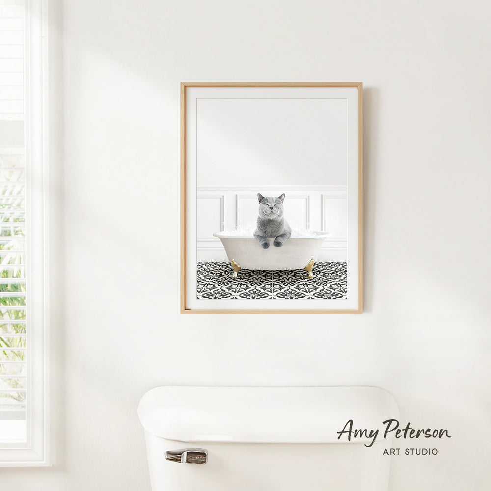 A framed image of a gray cat sitting in a white bathtub, with a patterned tile floor, is hanging on a white wall in a bathroom.