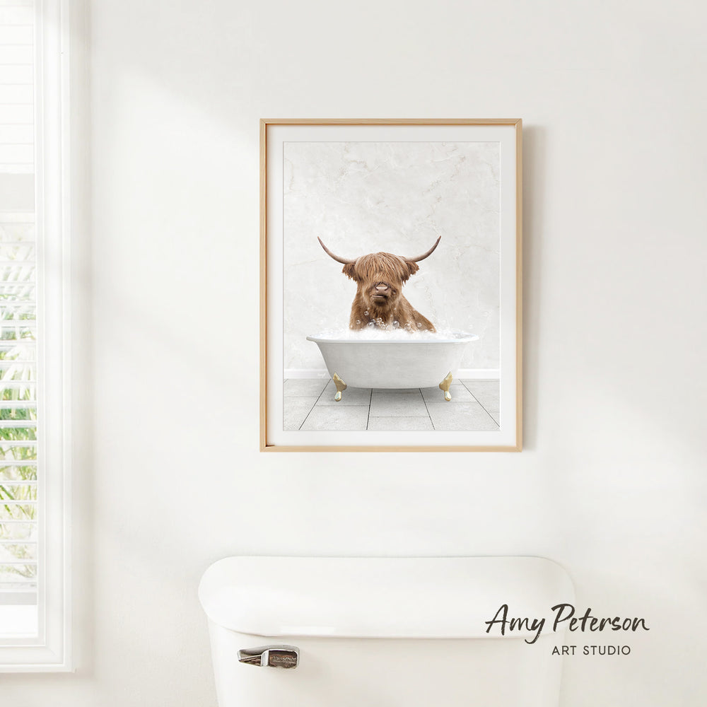 A framed image of a Highland cow sitting in a bathtub, with its horns raised, is hanging on a white wall.