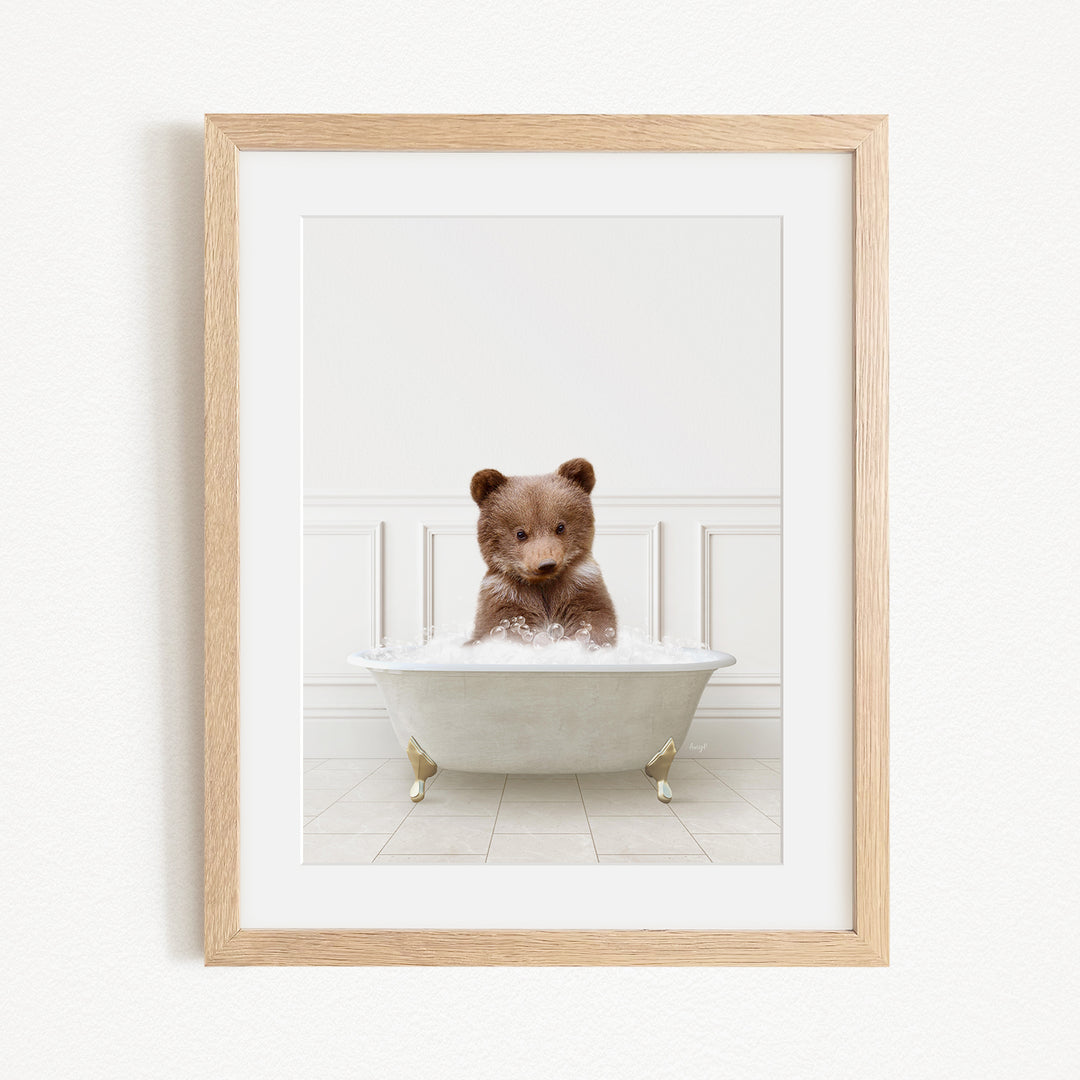 A cute brown bear cub sitting in a white bathtub filled with bubbles, looking directly at the camera.