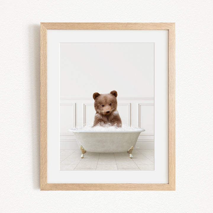 A cute brown bear cub sitting in a white bathtub filled with bubbles, looking directly at the camera.