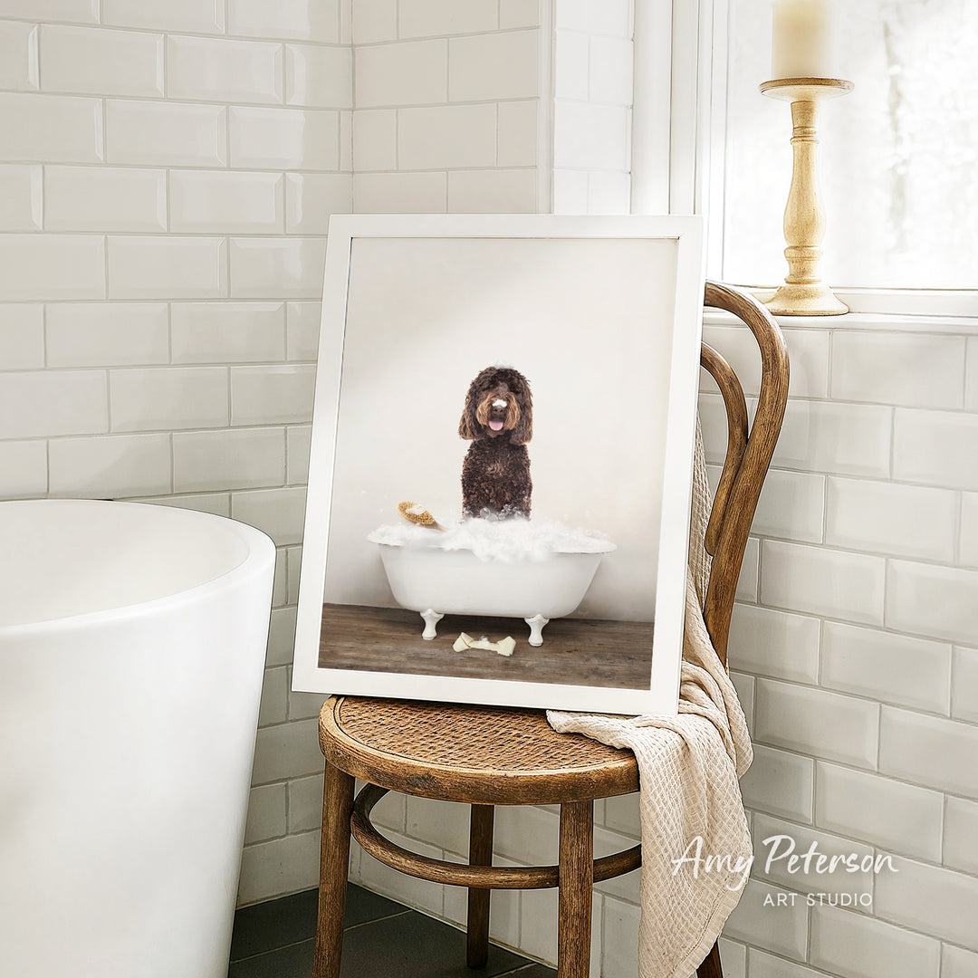 A framed photograph of a dog sitting in a bathtub, with a towel draped over the back of a wooden chair in a bathroom setting.
