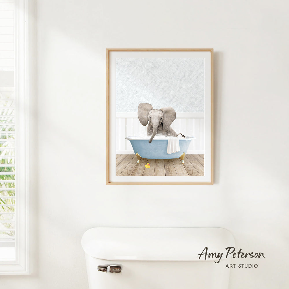 A framed print of an elephant sitting in a bathtub is hanging on a white wall.