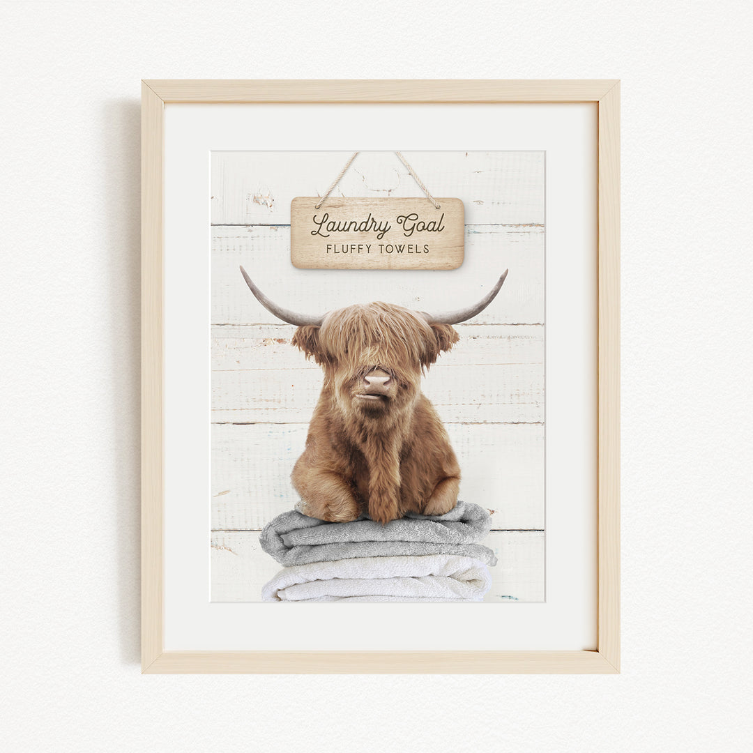 A framed image of a brown cow with long horns sitting on a stack of towels, with a sign above that says "Laundry Coal Fluffs Towels".