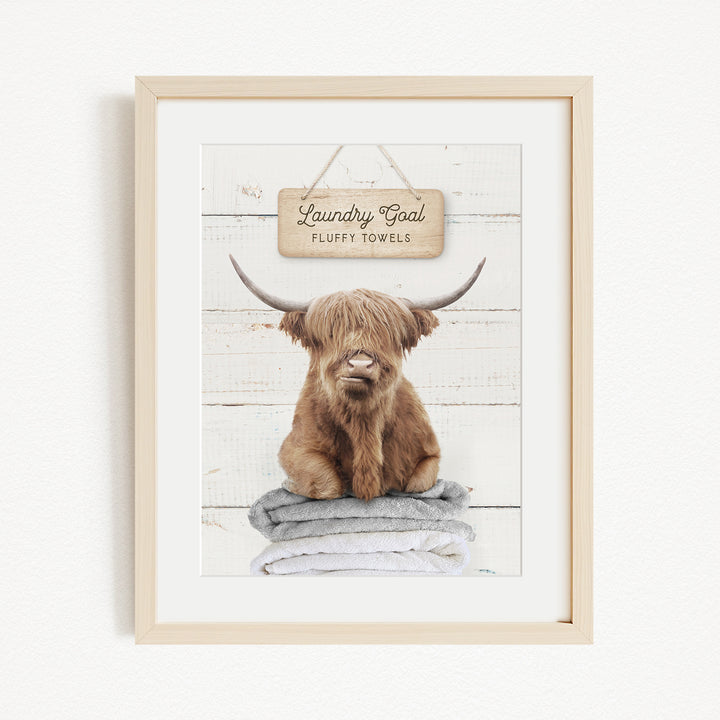A framed image of a brown cow with long horns sitting on a stack of towels, with a sign above that says "Laundry Coal Fluffs Towels".