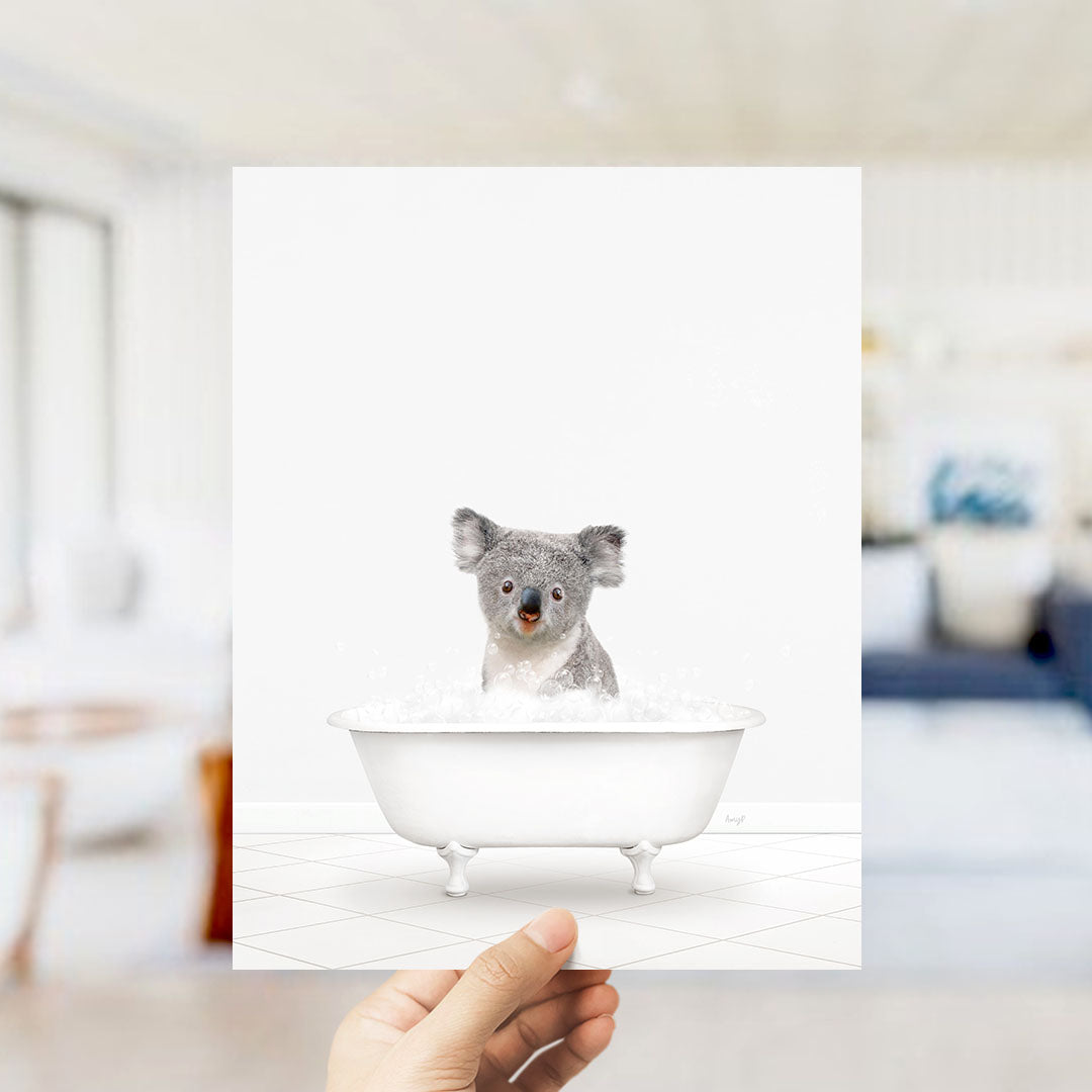 A hand holds up a framed image of a koala bear sitting in a bathtub filled with bubbles.