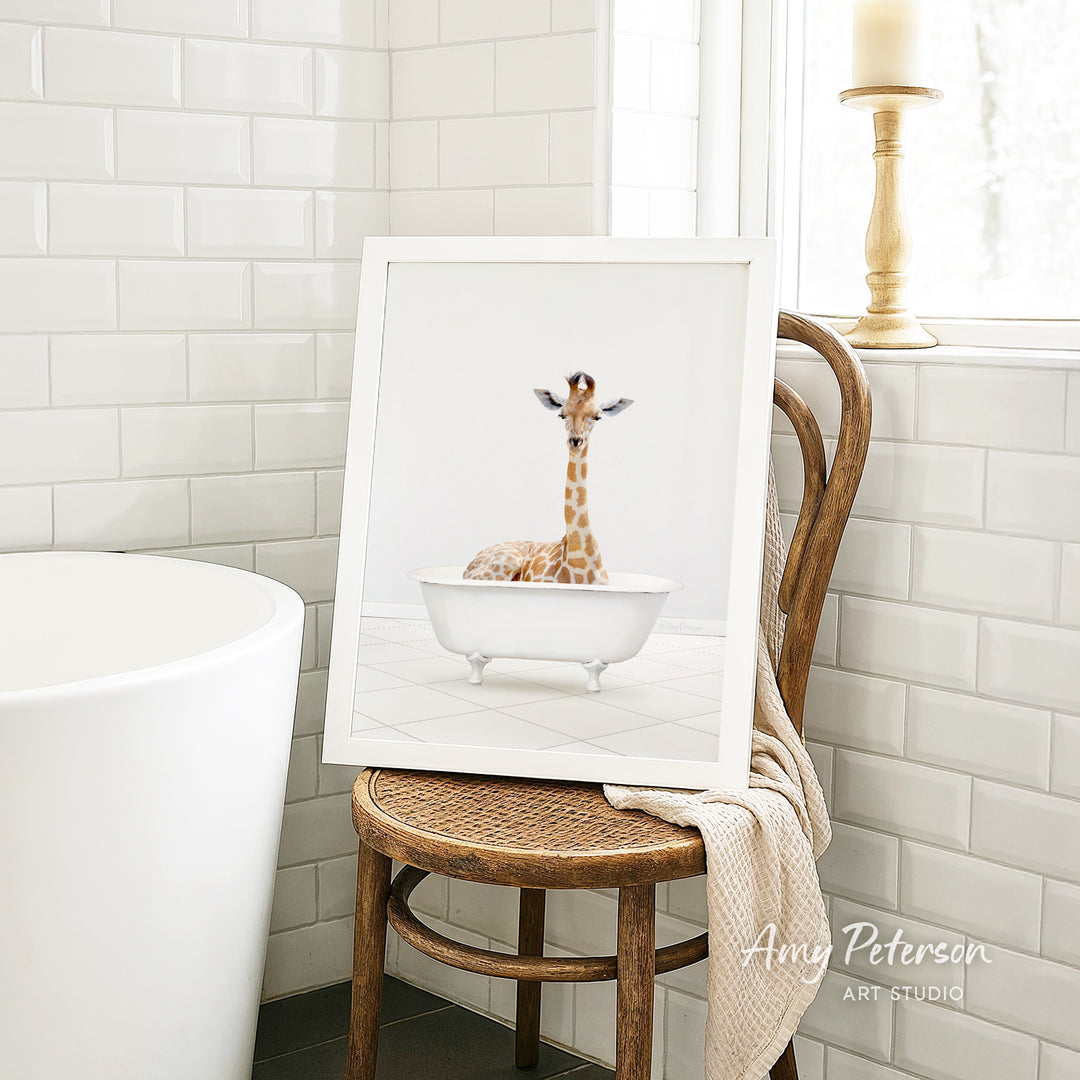 A framed image of a giraffe sitting in a white bathtub is displayed on a wooden chair in a bathroom setting.