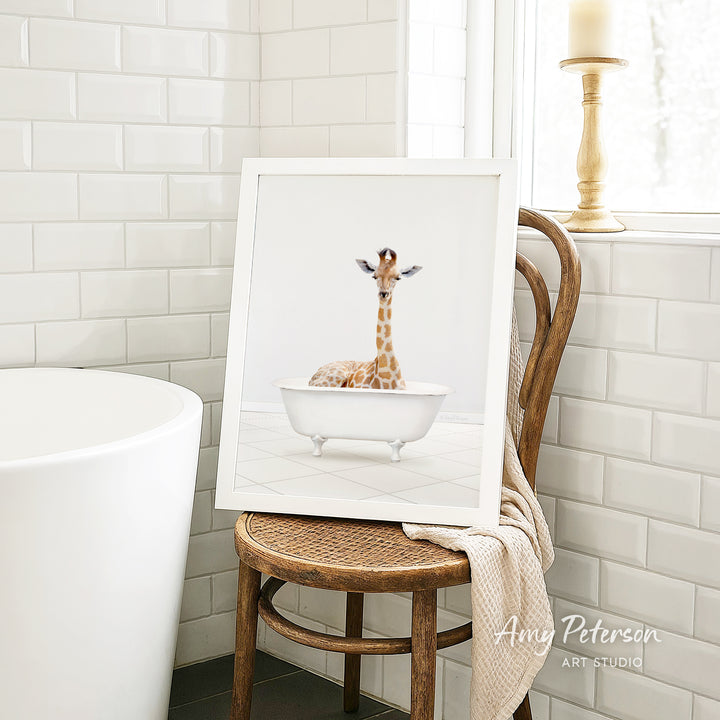 A framed image of a giraffe sitting in a white bathtub is displayed on a wooden chair in a bathroom setting.