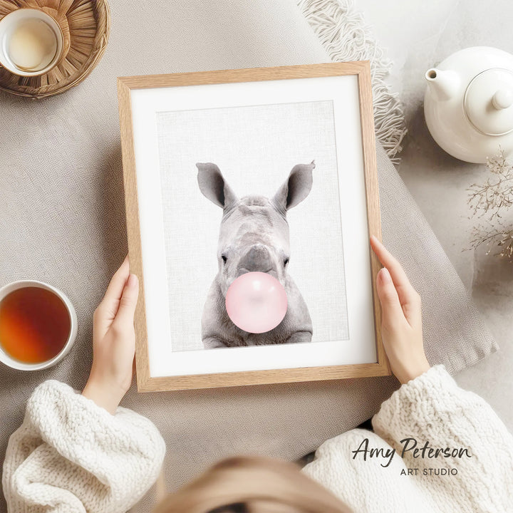 A person is holding a framed picture of a rhinoceros blowing a bubble with a pink bubblegum bubble.