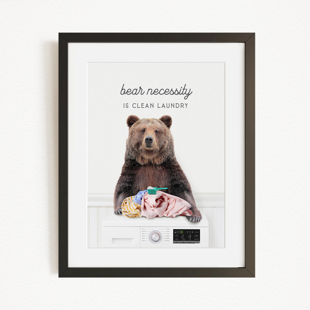 A framed image of a brown bear sitting on a washing machine, with the text "bear necessity is clean laundry" above it.