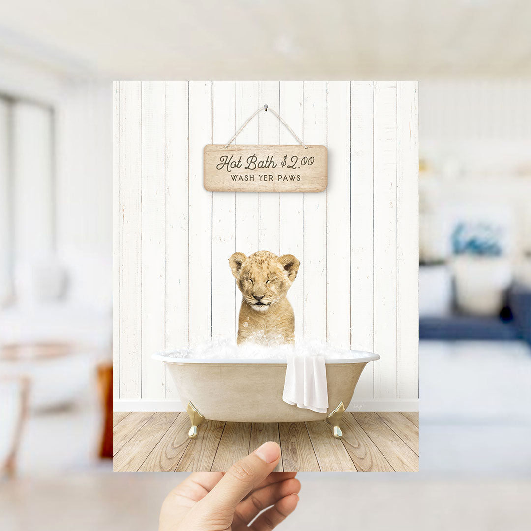 A hand holding a framed image of a young lion cub sitting in a bathtub, with a sign above the cub that reads "Hot Bath $2.00 WASH YOUR Paws".