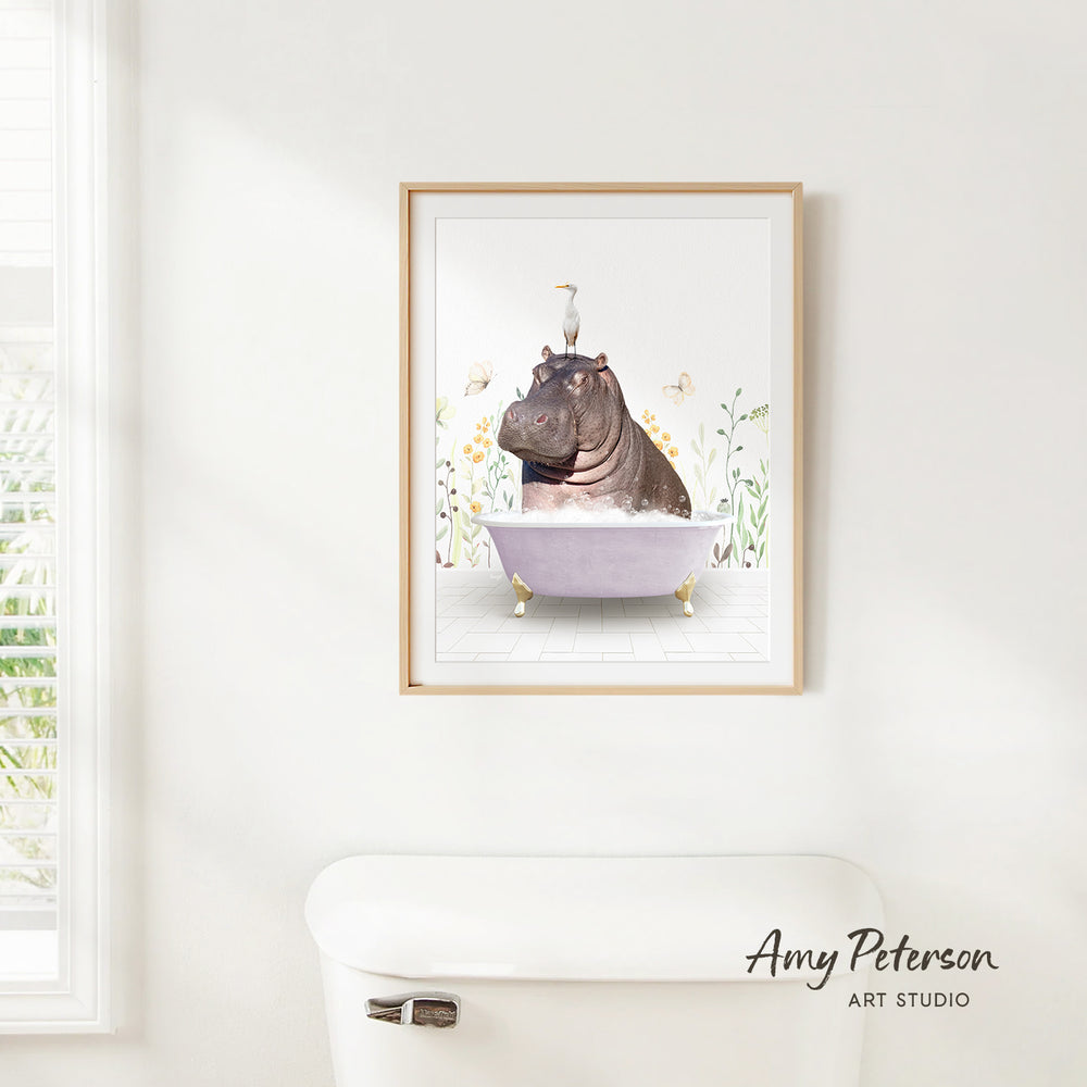 A framed illustration of a hippopotamus sitting in a bathtub, surrounded by flowers and butterflies, is hanging on a white wall.