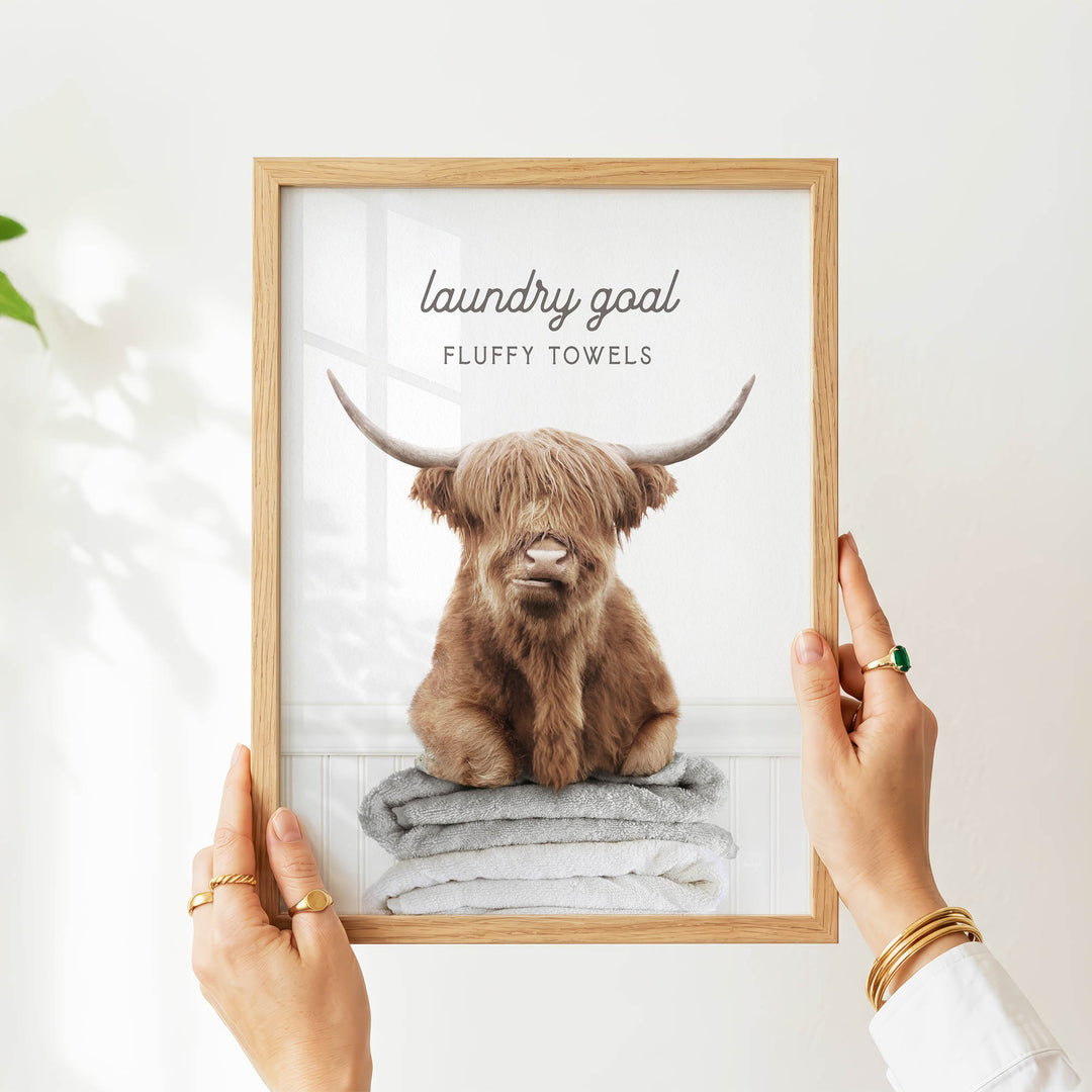 A person is holding up a framed picture of a brown cow with long horns, which is sitting on a stack of fluffy towels.